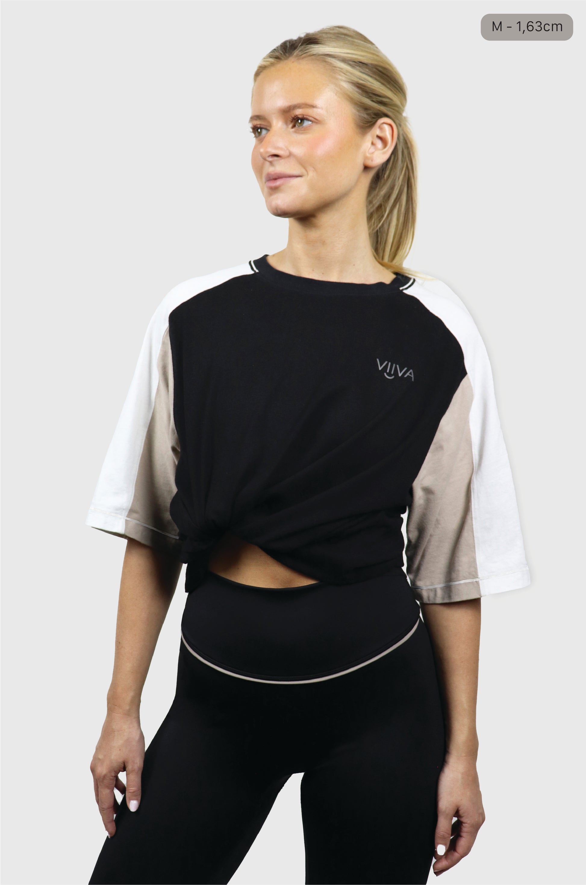 Woman wearing black, beige and white cotton t-shirt with grey small logo print at front print, raglan sleeves, black leggings on white background.