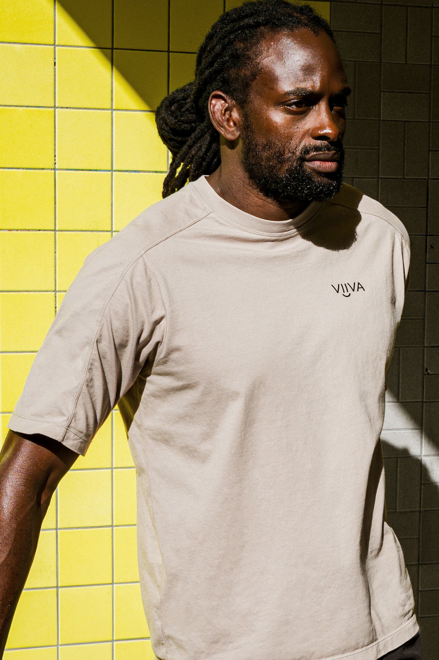 Man wearing a beige cotton t-shirt with 'VIIVA' logo against a yellow tiled wall