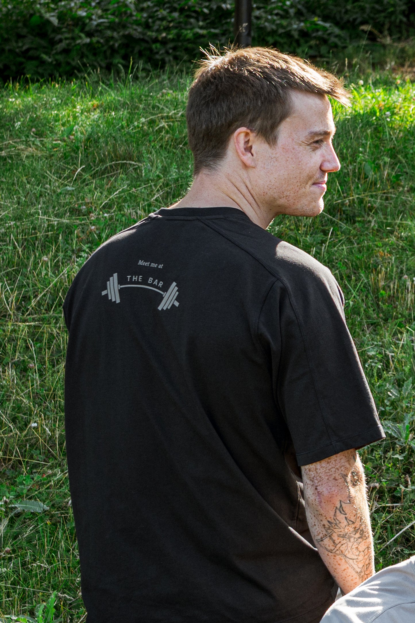 Man wearing a black cotton t-shirt with a print 'Meet me at the bar' on the back on a grass area