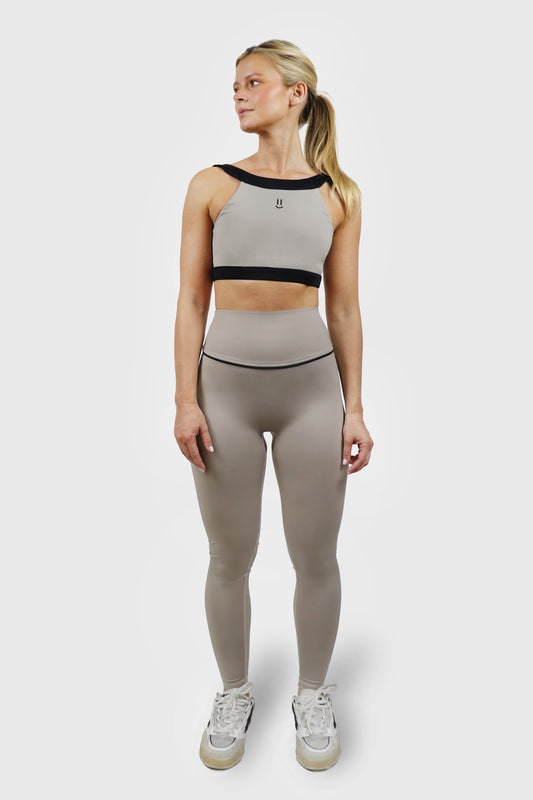 Woman wearing a beige taupe legging and top athletic outfit with black accents and details on a white background with small smiley print at front of top