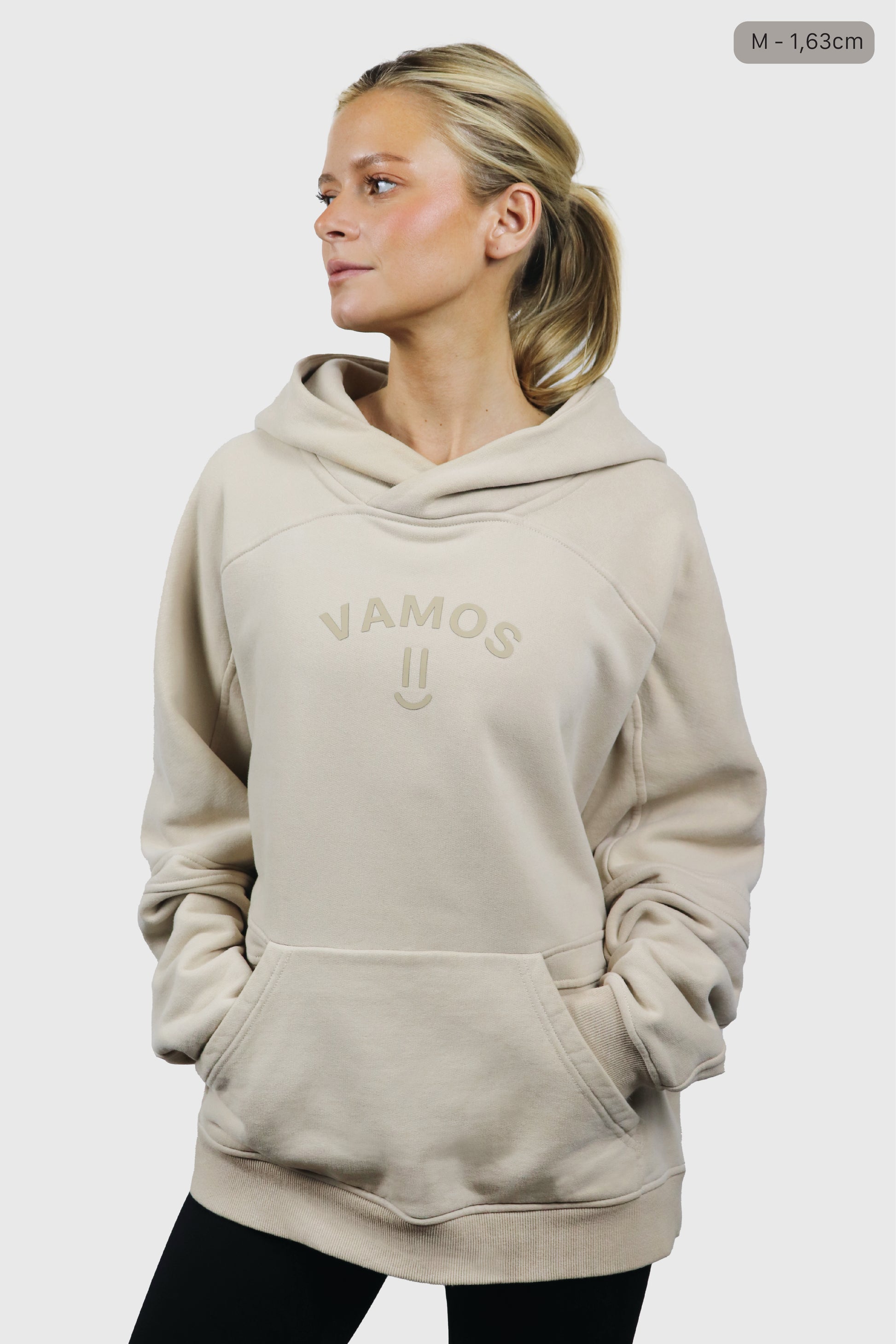 Woman wearing a beige cotton hoodie with print 'Vamos' at front with a smiley, and black leggings on a white background
