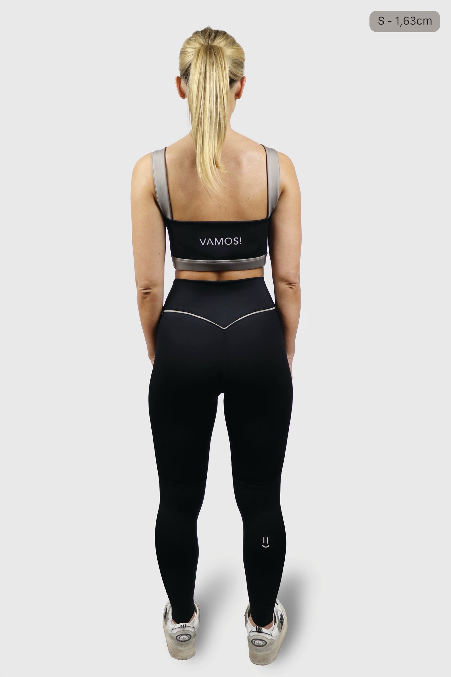 Woman wearing a black sports bra with 'VAMOS!' branding and taupe straps and black leggings with taupe details on a white background