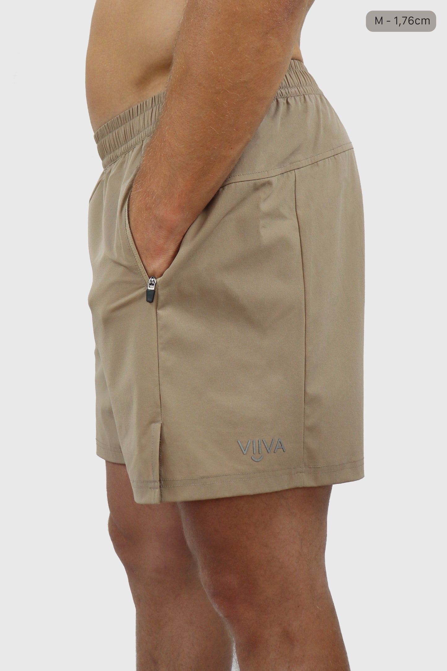 Man wearing beige shorts with a brand logo and hands in pocket on a plain background