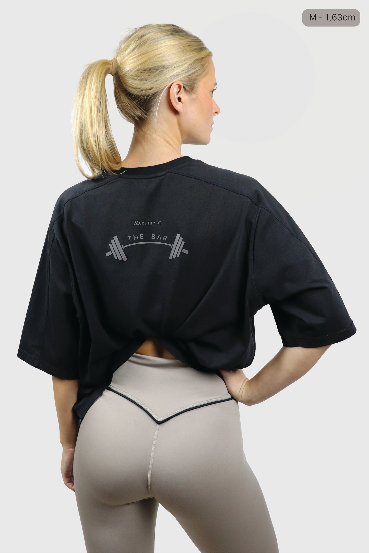 Woman wearing a black cotton t-shirt with a print 'Meet me at the bar' on the back against a white background