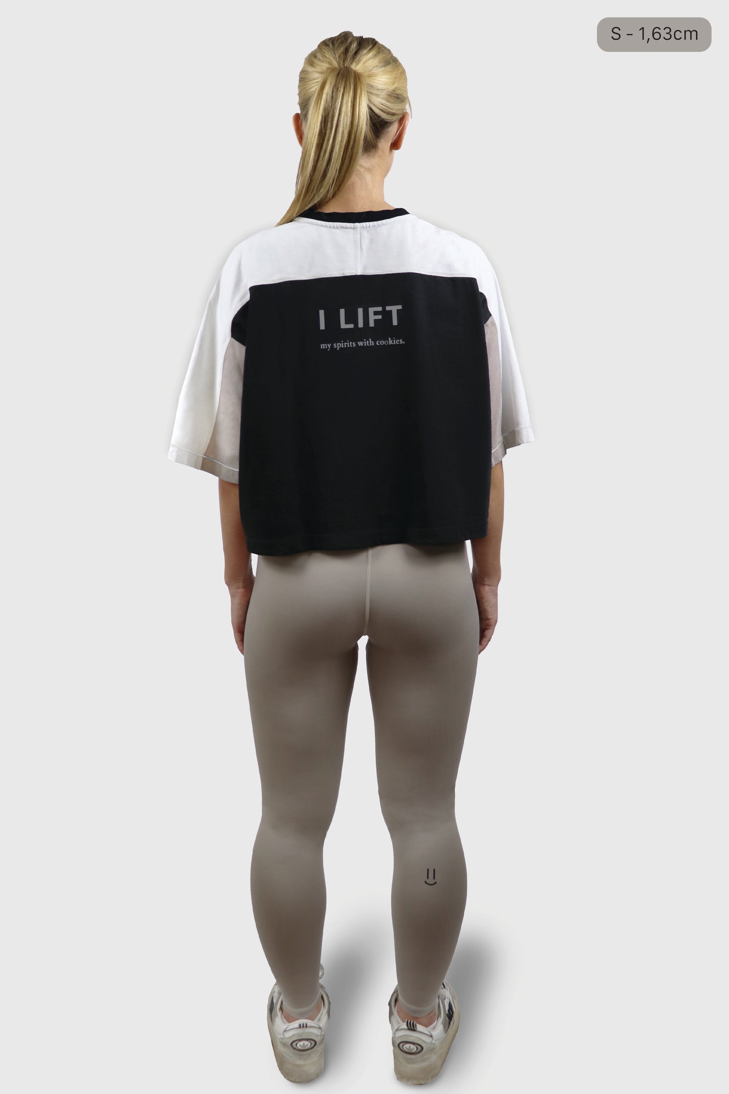 Woman wearing a black, beige and white cotton t-shirt with the phrase 'I LIFT my spirits with cookies' printed in grey at the back, and taupe leggings with a small smiley printed