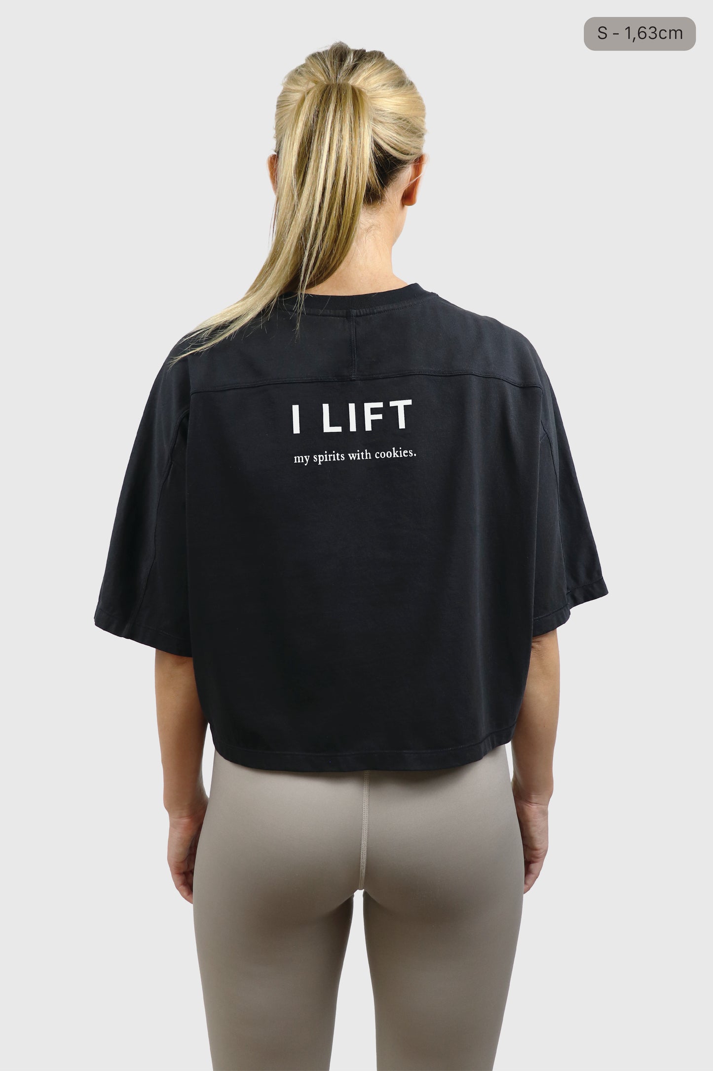 Woman wearing a black cotton t-shirt with 'I LIFT my spirits with cookies' text on the back, taupe leggings, standing against a plain background