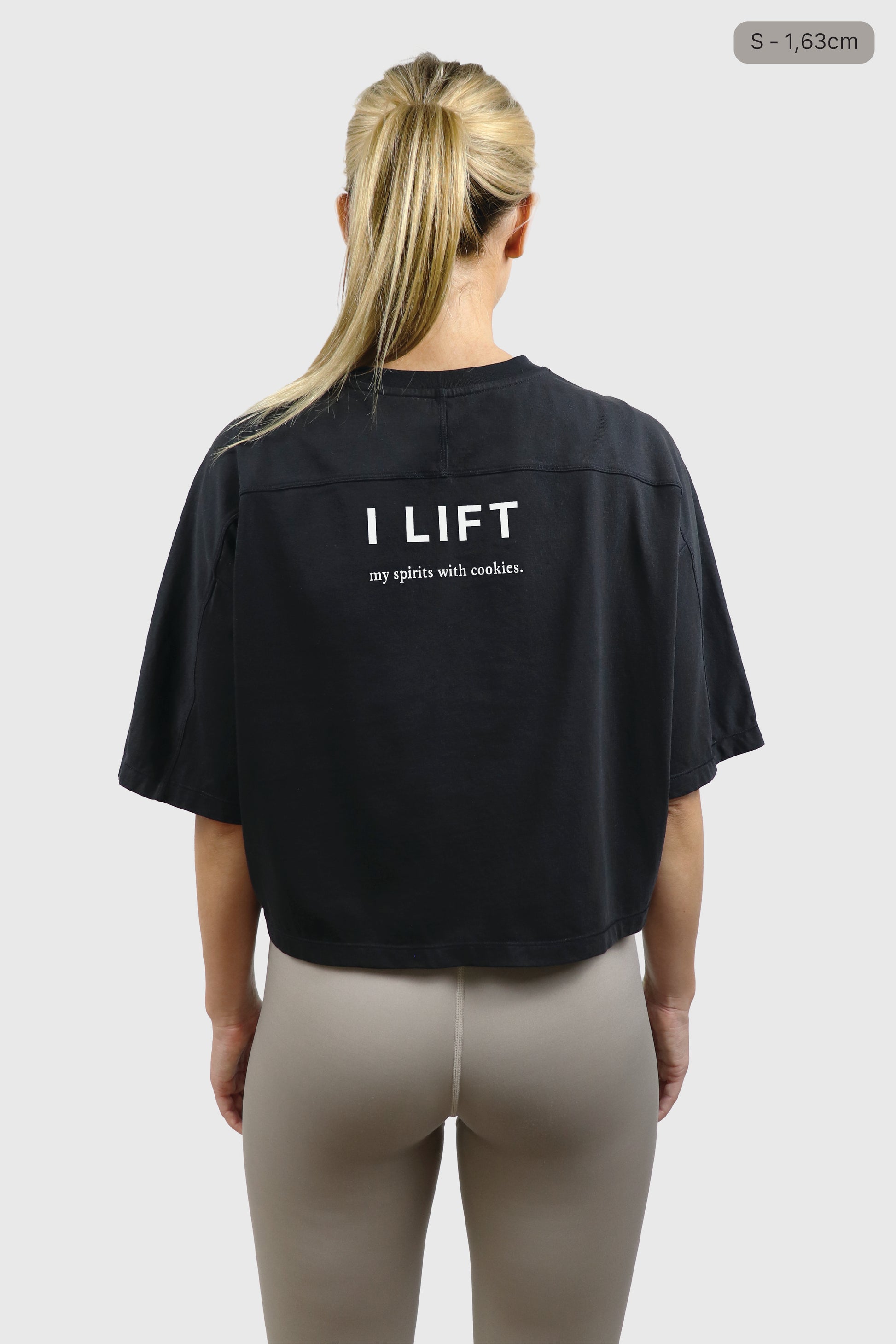 Woman wearing a black cotton t-shirt with 'I LIFT my spirits with cookies' text on the back, taupe leggings, standing against a plain background
