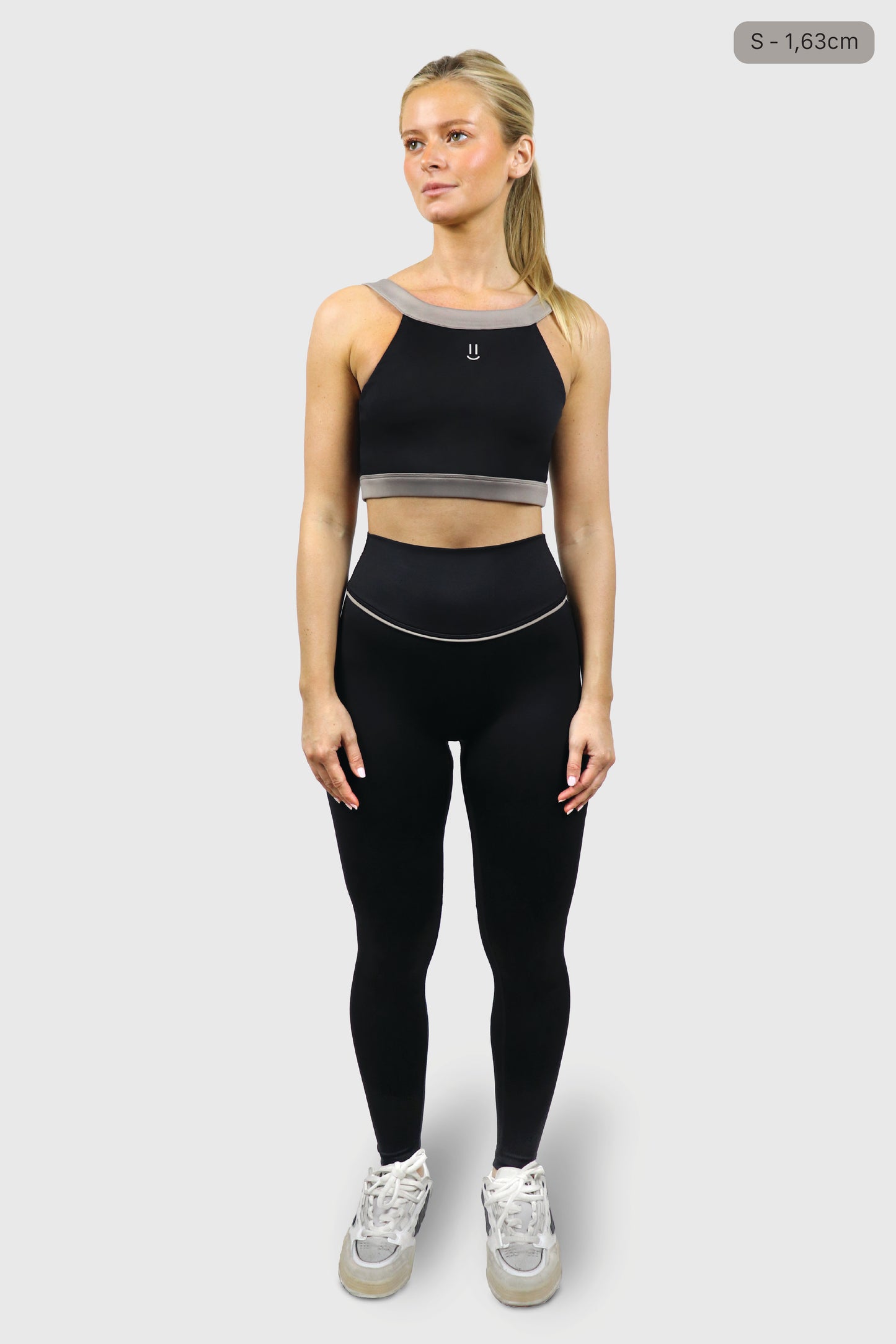 Woman wearing a black athletic top with small smiley print at front and leggings with gray taupe accents on a white background