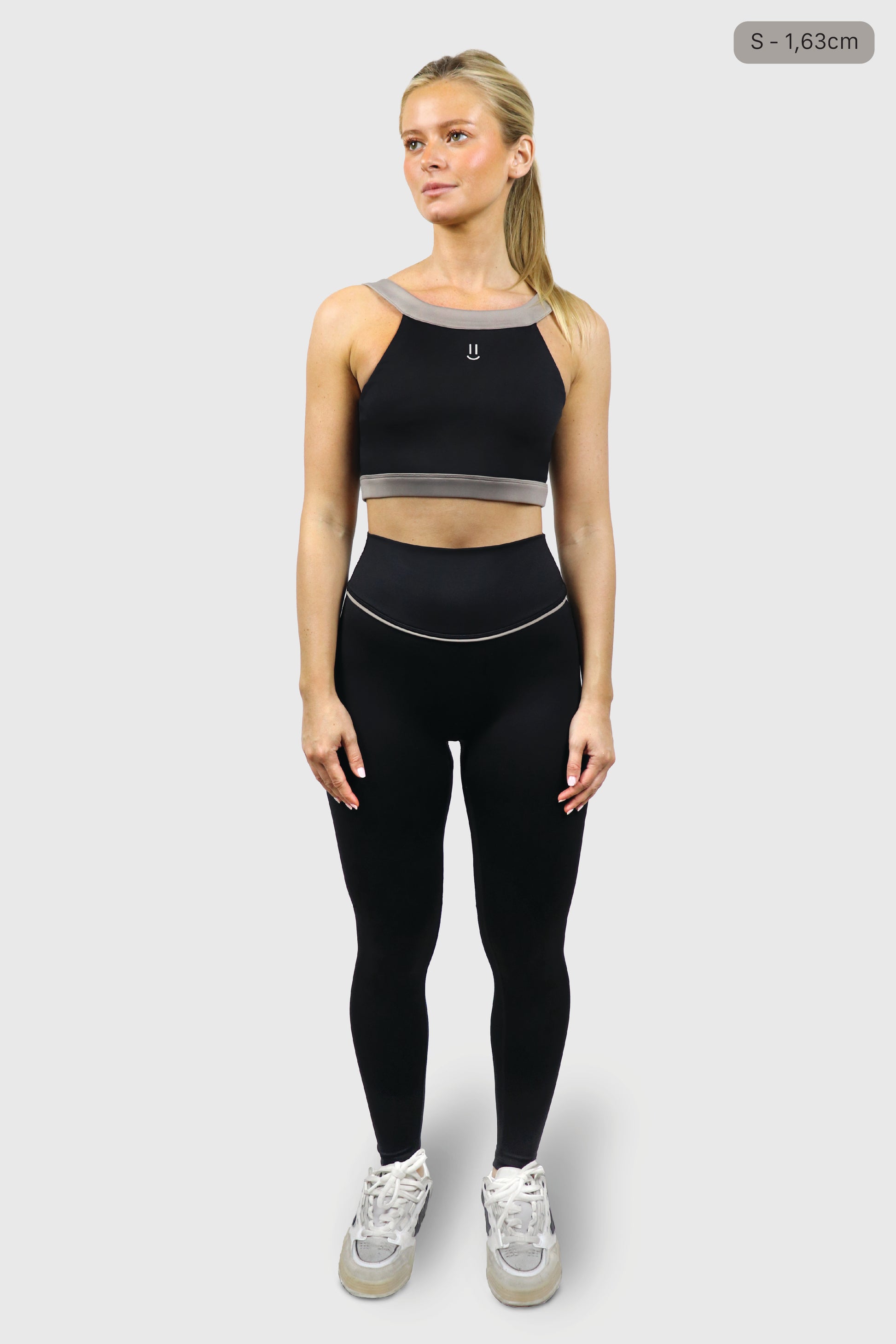 Woman wearing a black athletic top with small smiley print at front and leggings with gray taupe accents on a white background