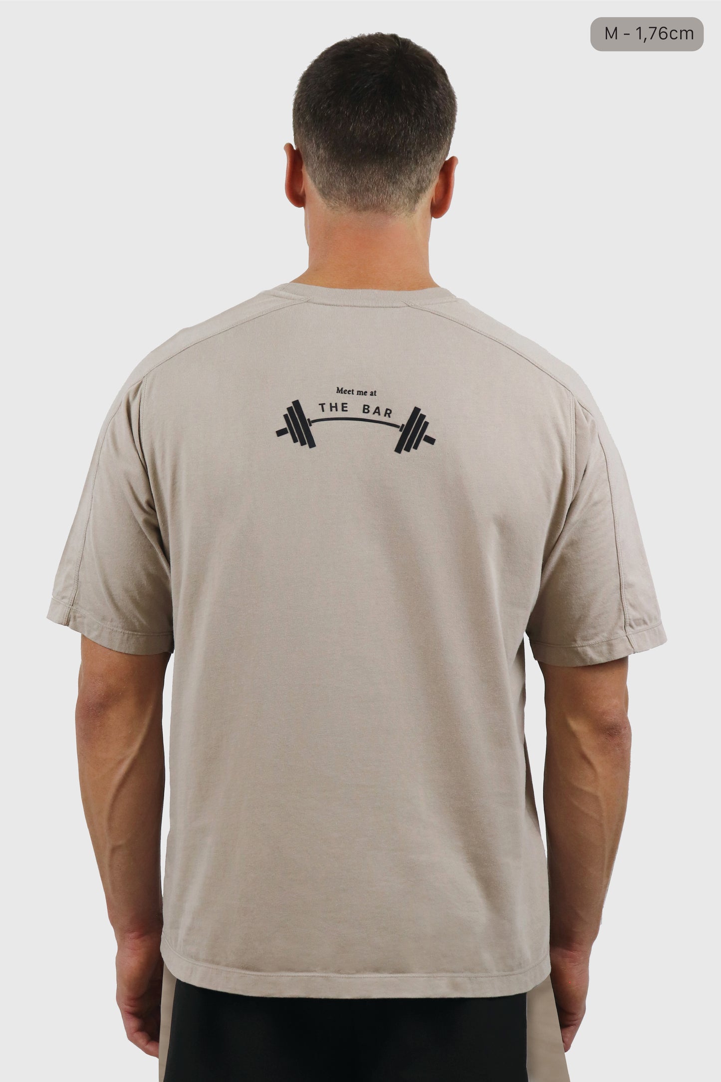 Man wearing a beige t-shirt with a print on the back written 'Meet me at the BAR' against a white background