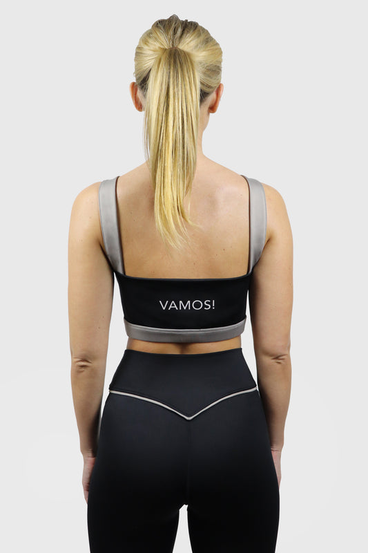 Woman wearing a black sports bra with 'VAMOS!' branding and taupe straps and black leggings with taupe details on a white background