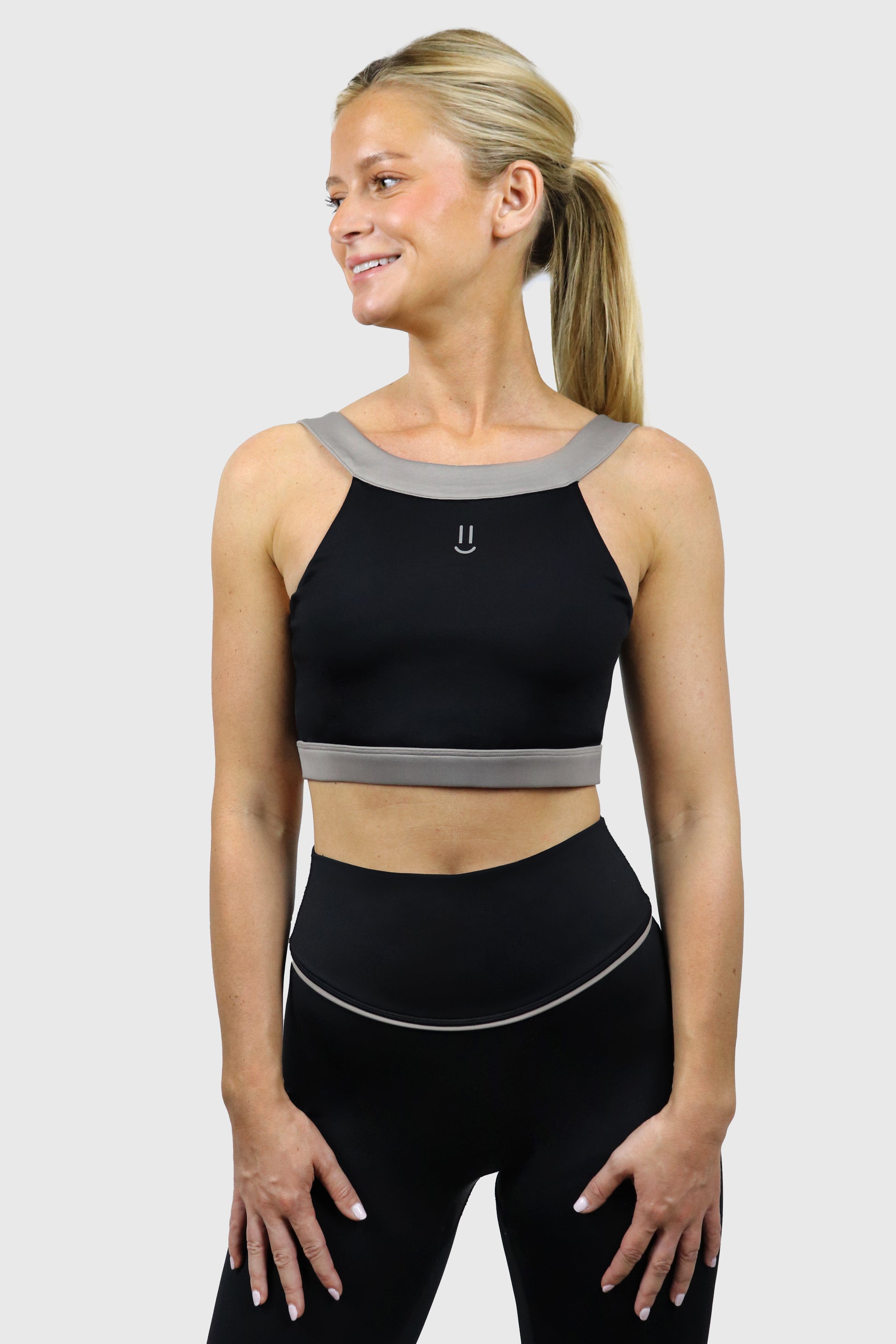Woman wearing a black athletic top with small smiley print at front and leggings with gray taupe accents on a white background
