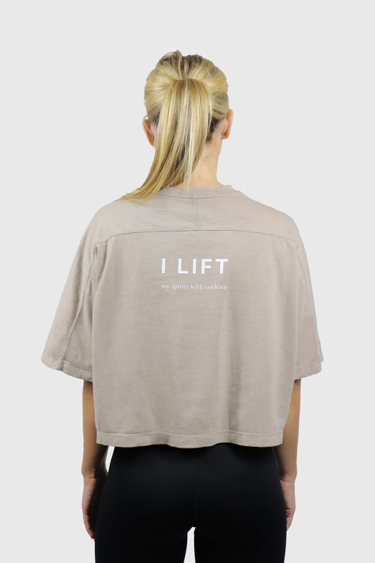 Woman wearing a beige cotton t-shirt with 'I LIFT my spirits with cookies' text on the back in white, standing against a white background