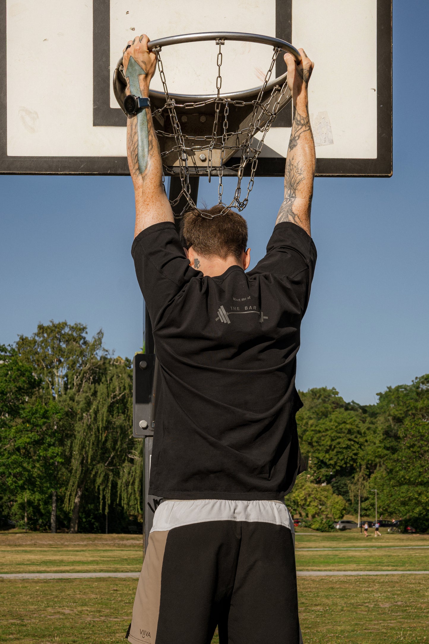 Man hanging from a basketball hoop in an outdoor setting with trees and clear sky wearing cotton black t-shirt with 'bar' printand color block shorts