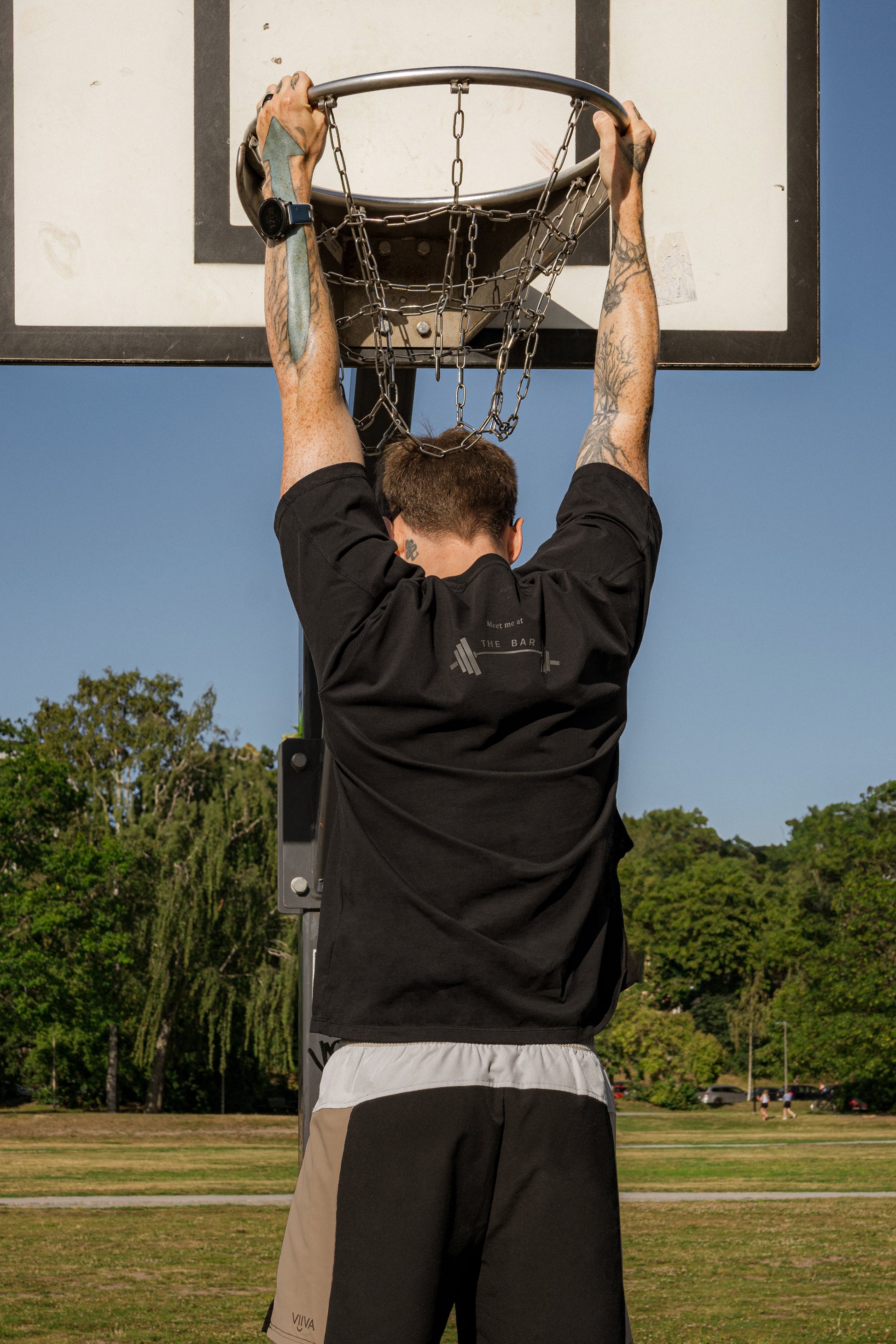 Man hanging from a basketball hoop in an outdoor setting with trees and clear sky wearing cotton black t-shirt with 'bar' printand color block shorts