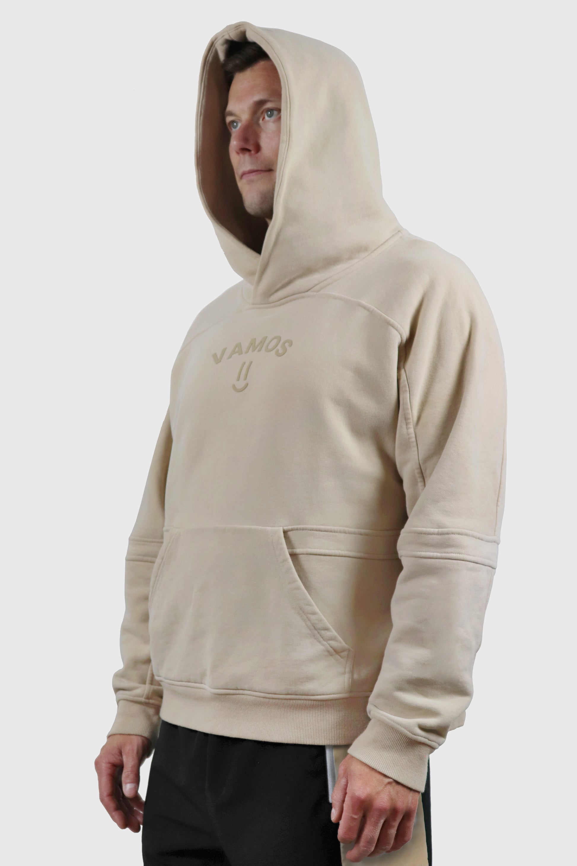 Man wearing a beige hoodie with 'Vamos' print at front with a smiley on a white background