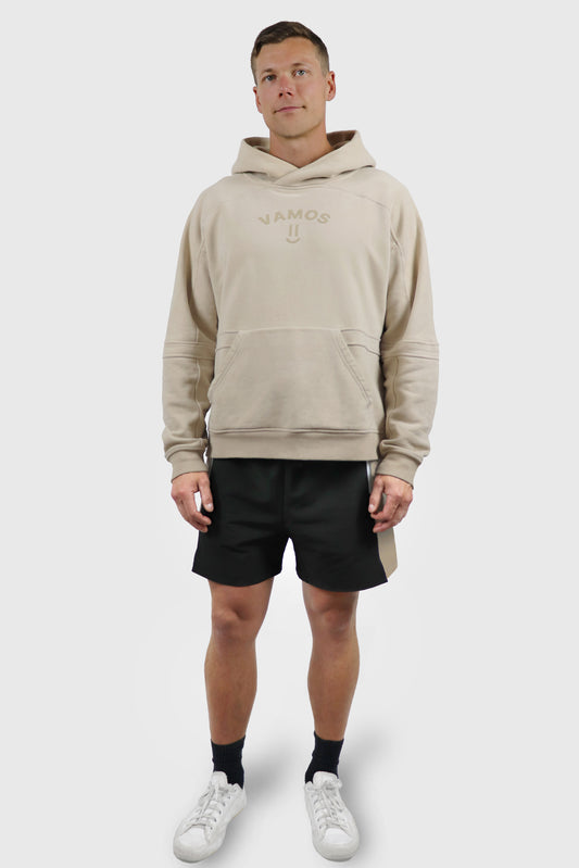 Man wearing a beige cotton hoodie with print 'Vamos' at front with a smiley, and black shorts on a white background