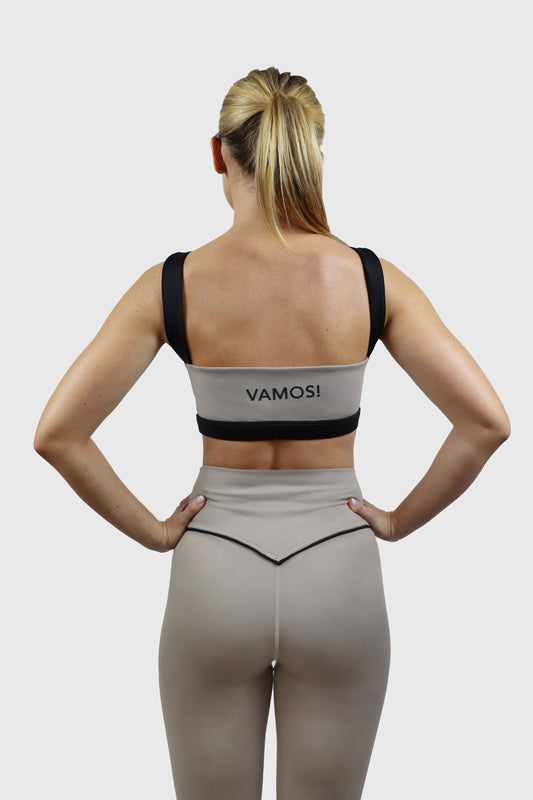 Woman wearing a beige taupe sports bra with black straps and 'vamos!' print at back and taupe leggings with black accents on a white background