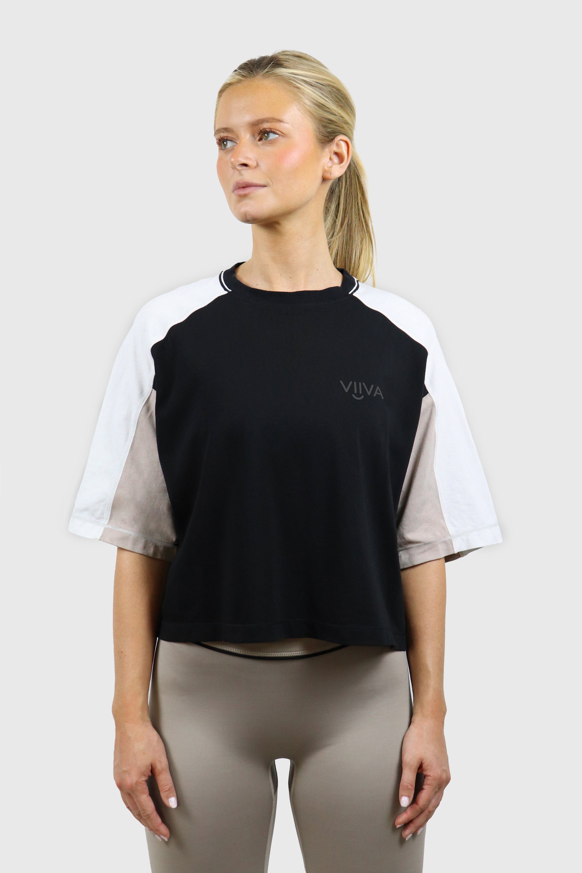 Woman wearing a black, beige and white cotton t-shirt with raglan sleeves and small logo print, and taupe leggings with black piping detail