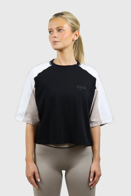 Woman wearing a black, beige and white cotton t-shirt with raglan sleeves and small logo print, and taupe leggings with black piping detail