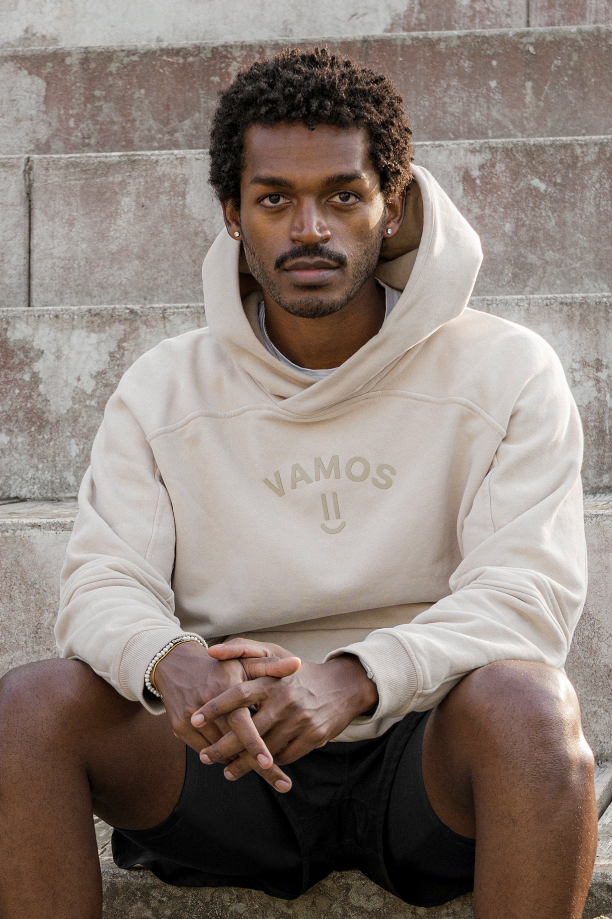 Man wearing a beige hoodie with 'Vamos' logo  with a smiley sitting against a stone wall.