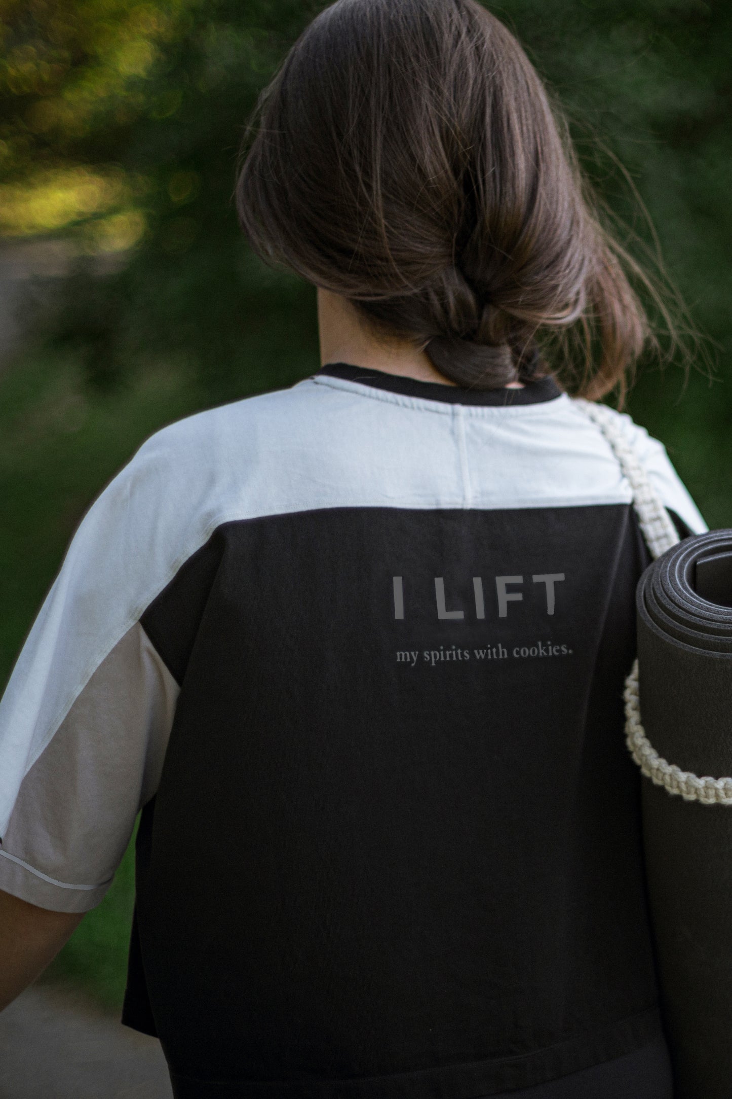 Person wearing a cotton t-shirt in white, black and beige with 'I LIFT my spirits with cookies' text, holding a yoga mat outdoors.