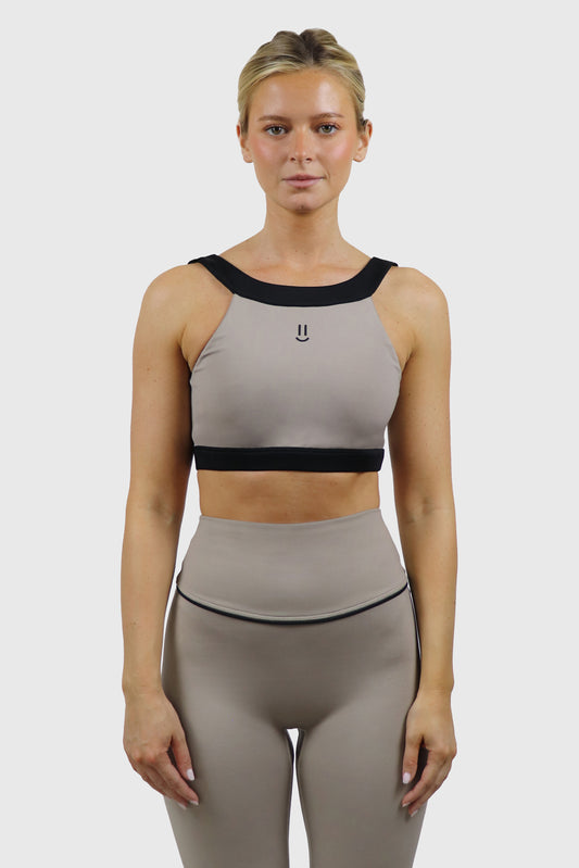 Woman wearing a beige taupe sports bra and taupe leggings with black accents on a white background