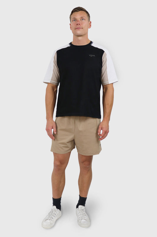 Man wearing cotton black, beige and white t-shirt with raglan sleeves, grey small logo print at front, beige shorts, white background.