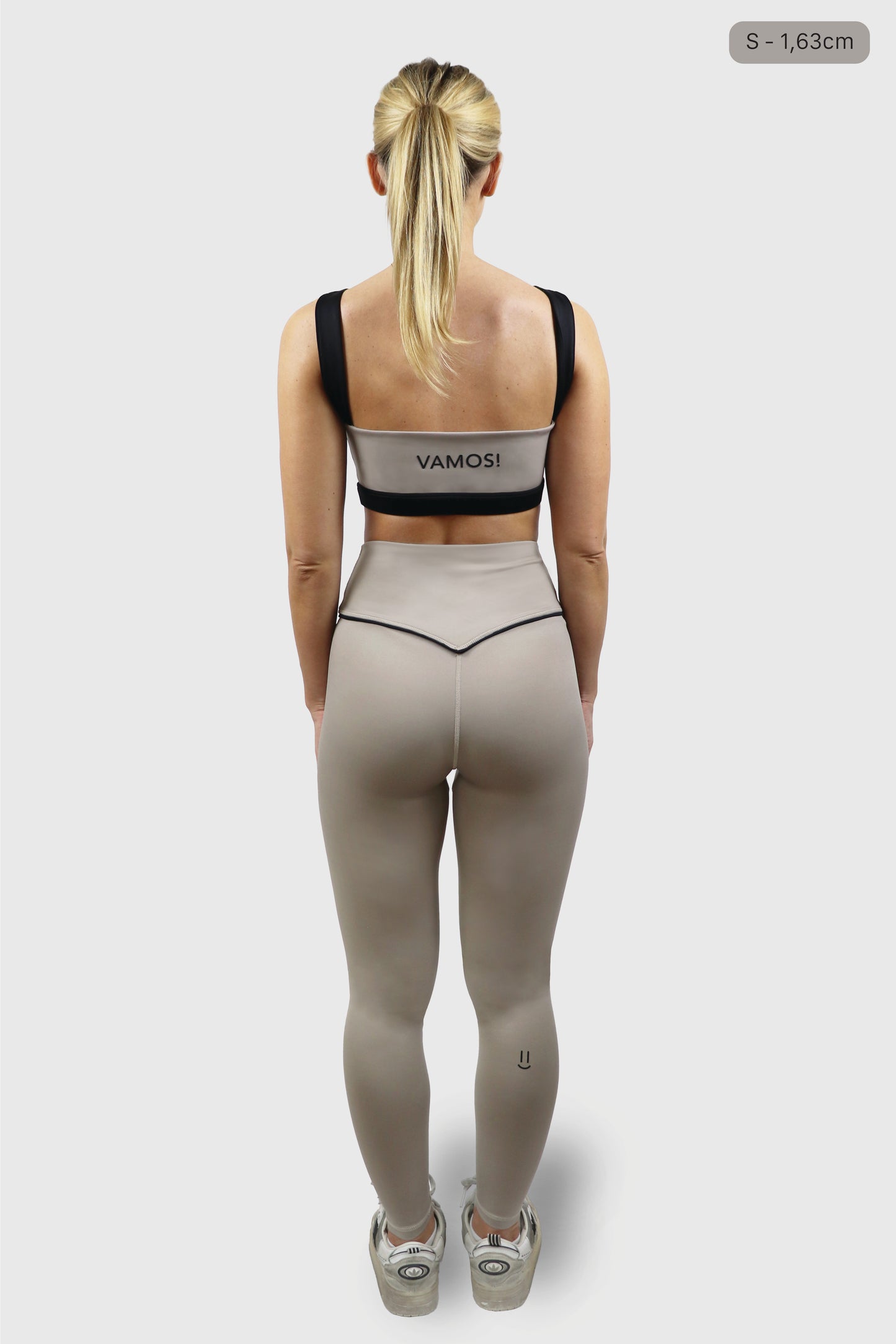 Woman wearing beige/ taupe sports bra with black straps with print of the word VAMOS and beige/ taupe leggings with black detail