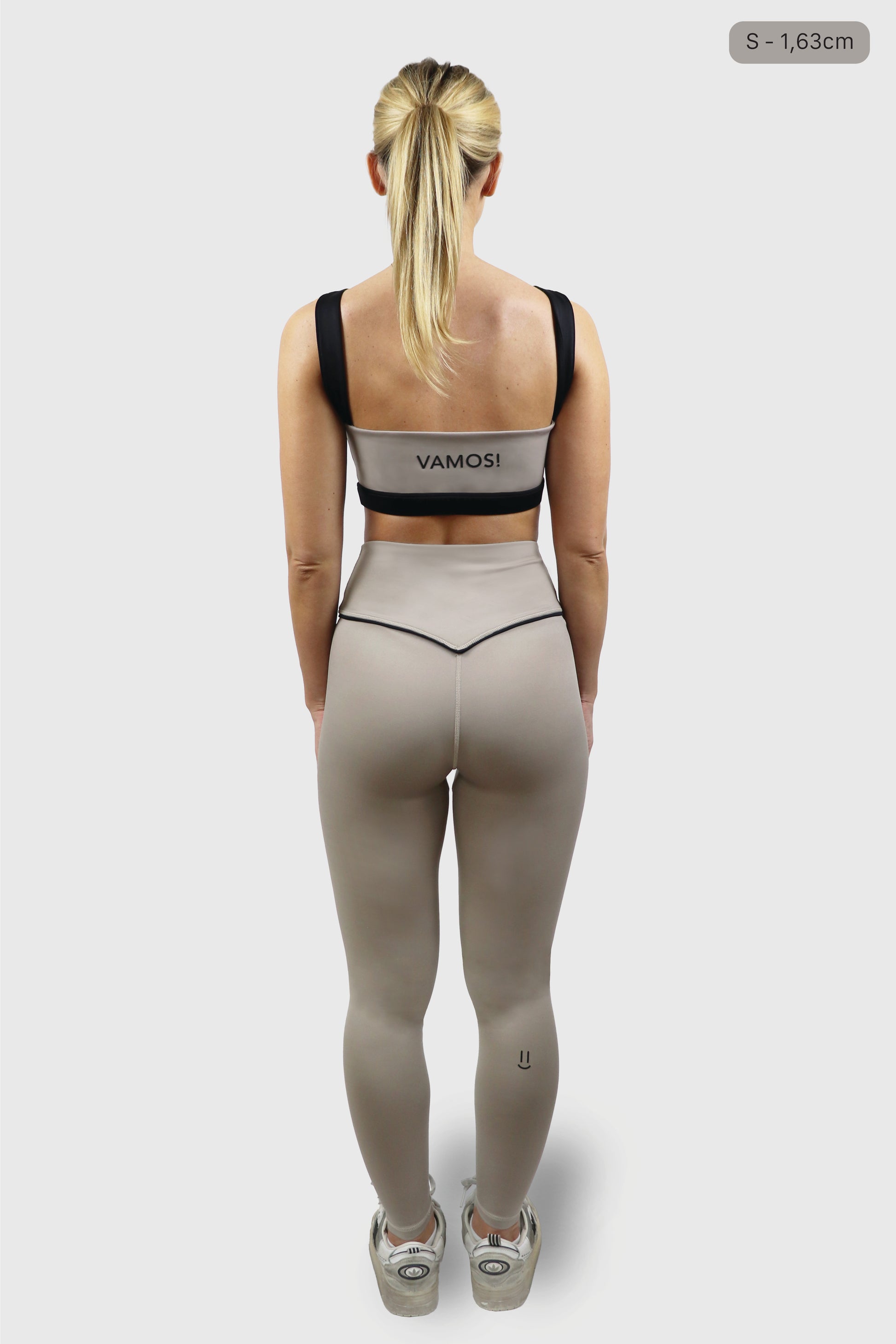 Woman wearing beige/ taupe sports bra with black straps with print of the word VAMOS and beige/ taupe leggings with black detail