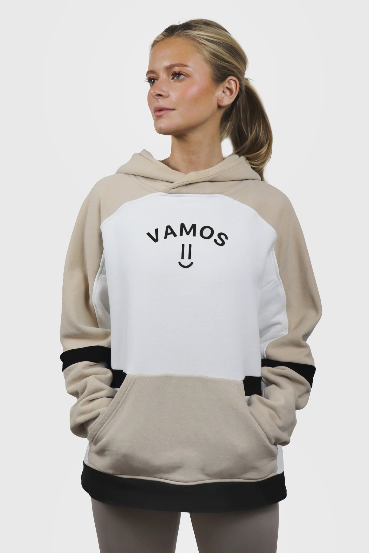 Woman wearing a beige and white hoodie with 'VAMOS' print in black at front wion a plain backgroundh smiley printed as well, and black stripes details